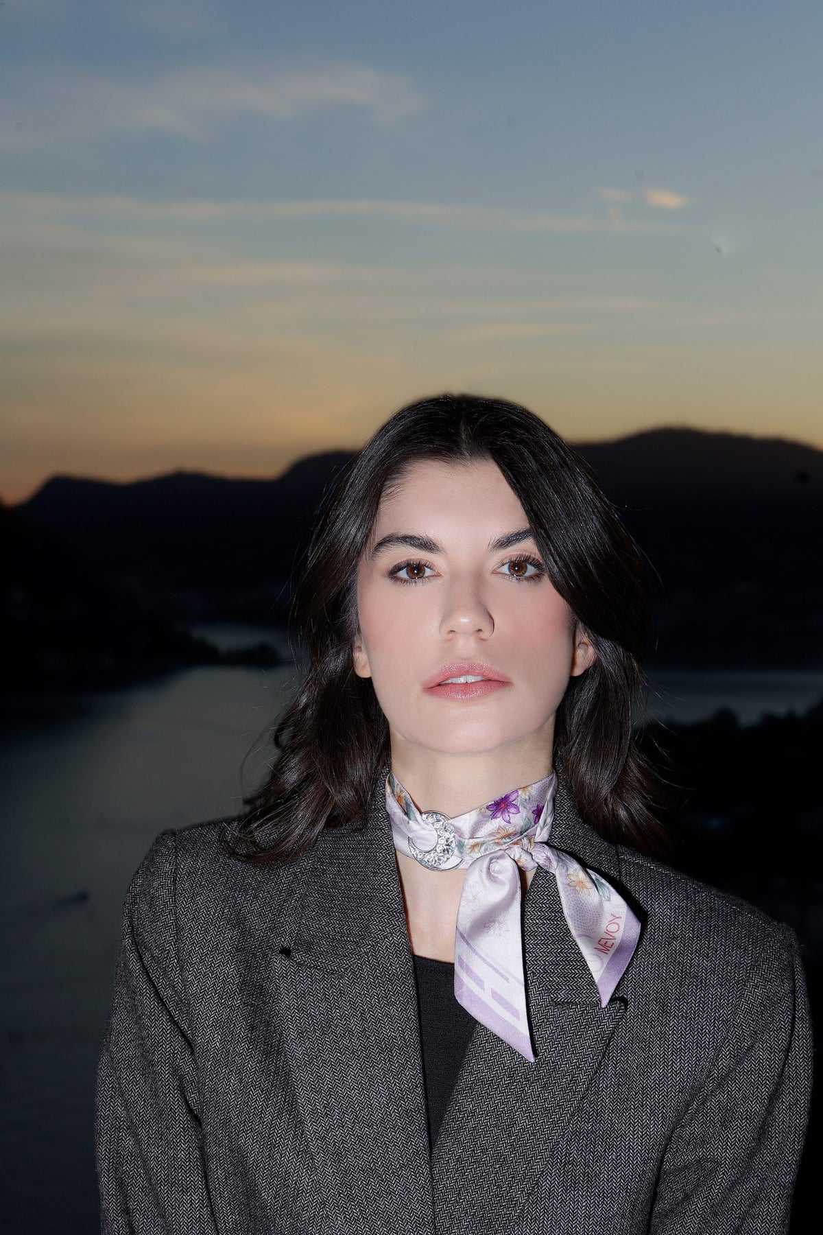 Frau im grauen Blazer mit einem fliederfarbenen MEVOY MeSilk am Hals, hinter ihr der Comer See bei Sonnenuntergang