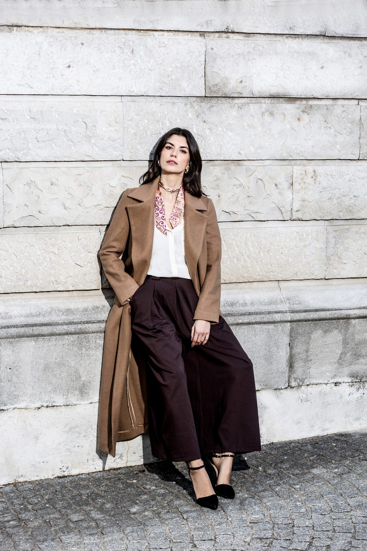 Frau im langen Kamelhaarmantel mit einem MEVOY MeSilk am Hals, an die Steinmauer einer Villa in Como gelehnt